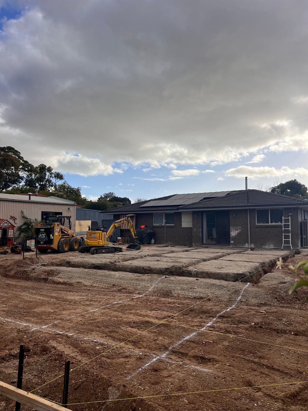 Construction site with yellow excavator, modern dark building, and dirt foundation work under cloudy sky