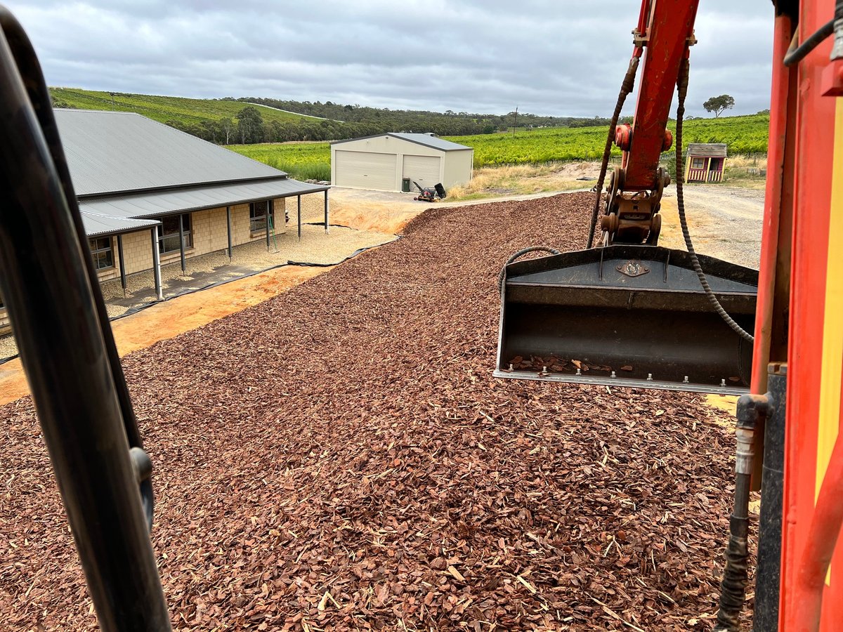 Red excavator with bucket in foreground over gravel yard at farm with buildings and green fields in background