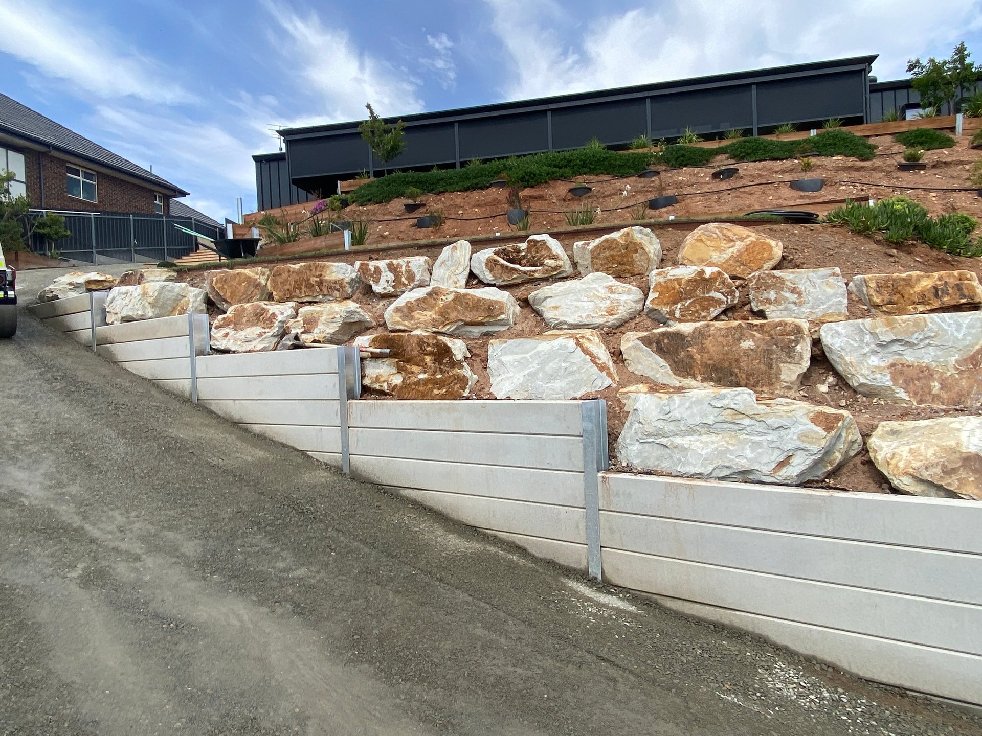 Large tan and brown boulders stacked against a white retaining wall with landscaping above and buildings in the background