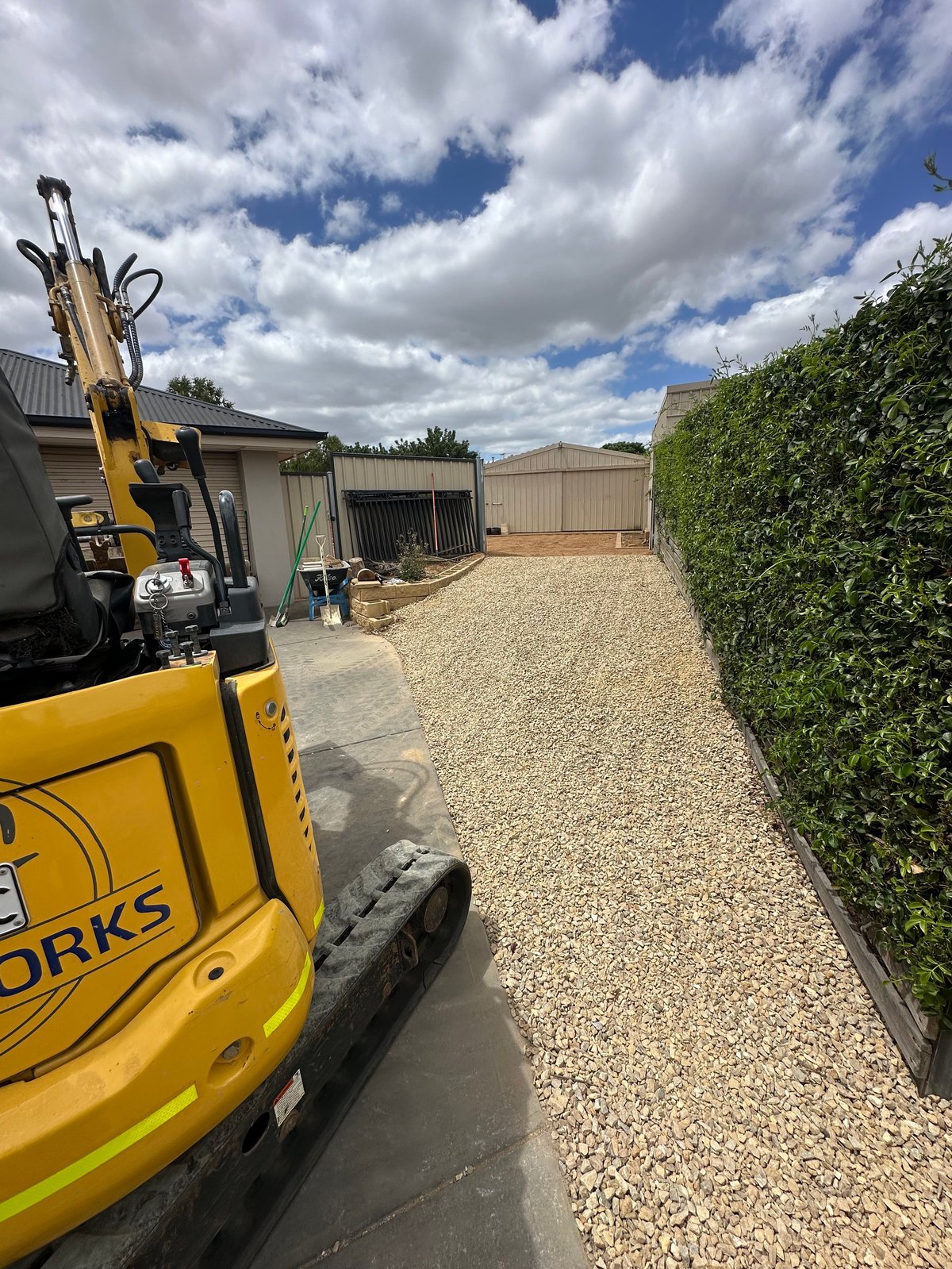Yellow mini excavator parked on gravel pathway beside industrial buildings under partly cloudy sky