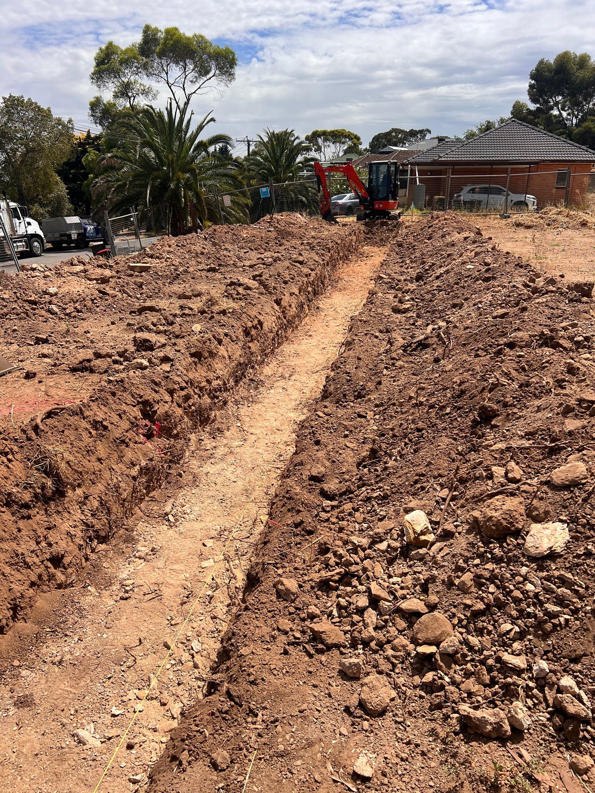 Excavated trenches in brown soil on a construction site with brick house, palm trees, and heavy machinery visible in background