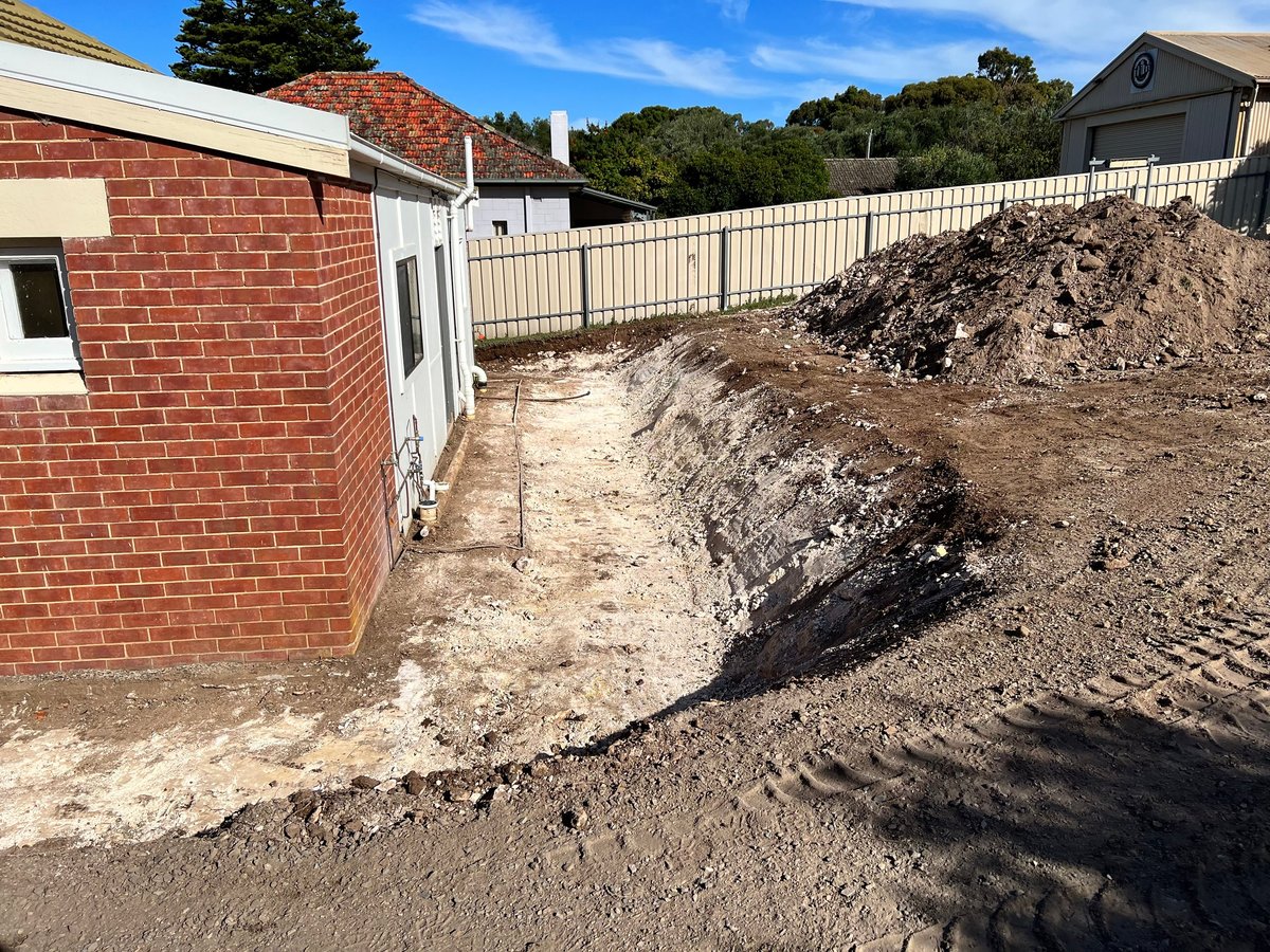 Construction site with red brick building, metal fence, piles of dirt and gravel, surrounded by trees and blue sky