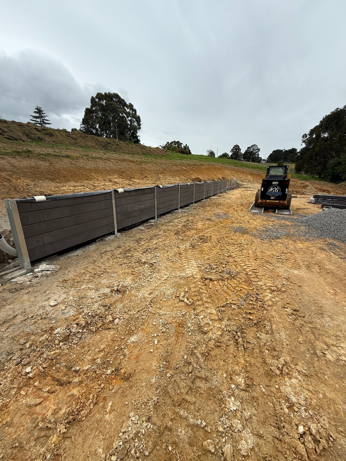 Construction site with dark wooden fence, gravel driveway, and small loader vehicle on a hillside under cloudy sky
