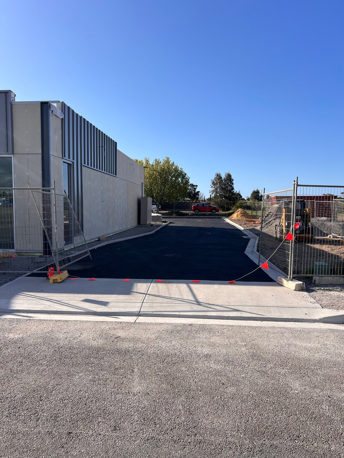 Construction site with freshly poured black asphalt driveway, safety barriers, industrial buildings, and clear blue sky
