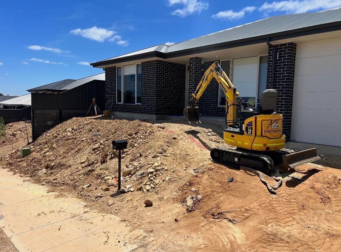 Yellow mini excavator on construction site with newly built brick house and dirt yard under clear blue sky
