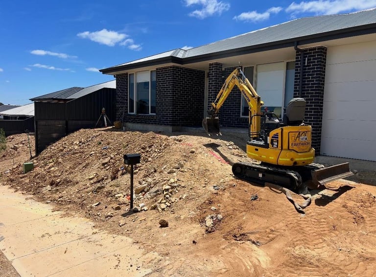 Yellow mini excavator on construction site with newly built brick house and dirt yard under clear blue sky