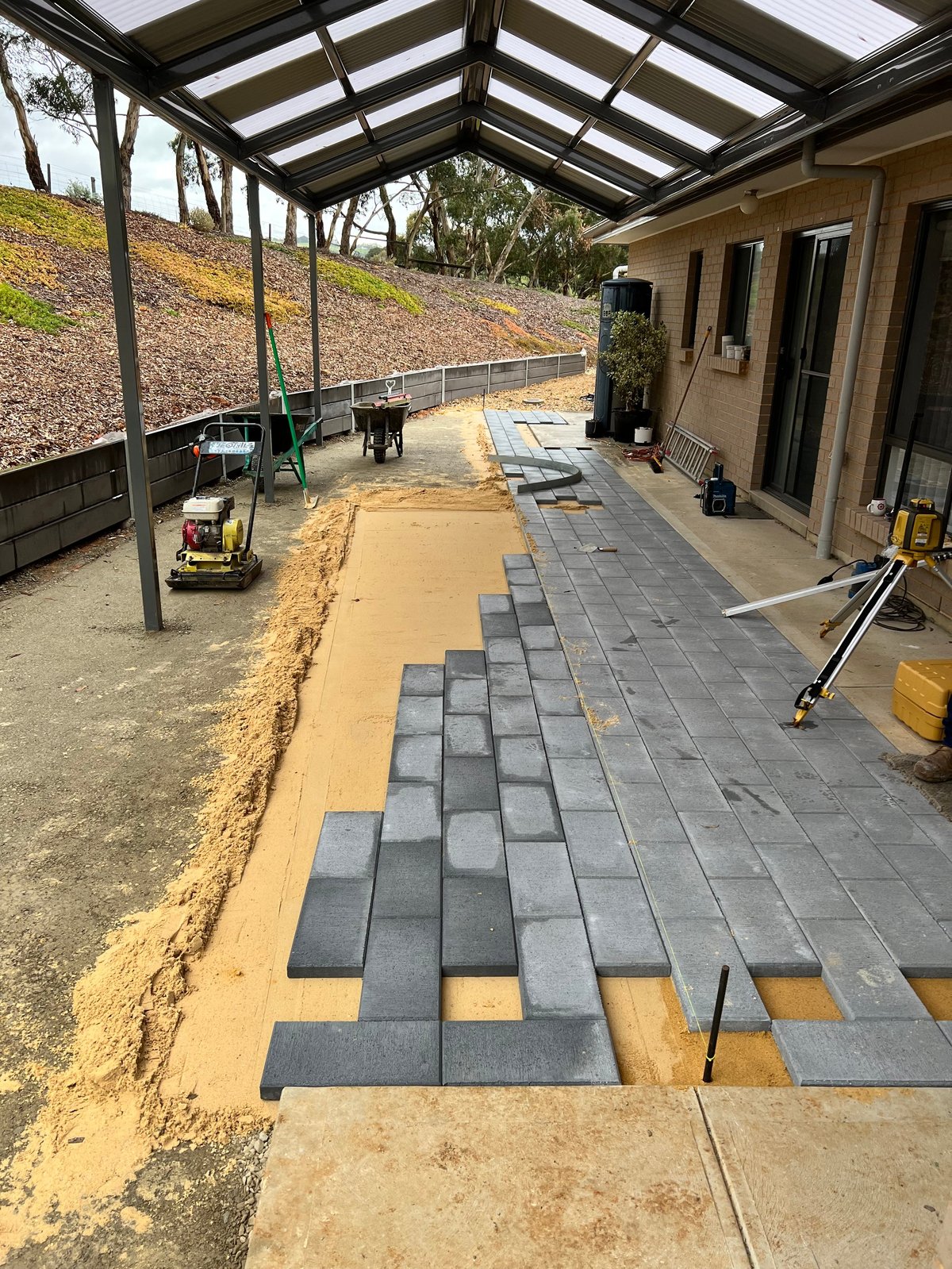 Patio installation in progress with dark pavers being laid on sand base under a pergola structure attached to a tan-colored house