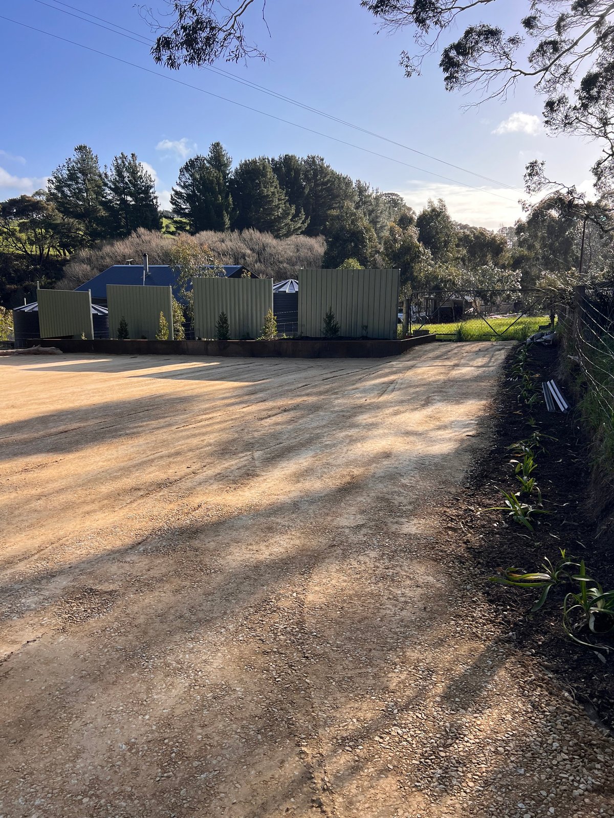 Rural property with corrugated metal storage buildings on unpaved driveway surrounded by trees and green fields under blue sky