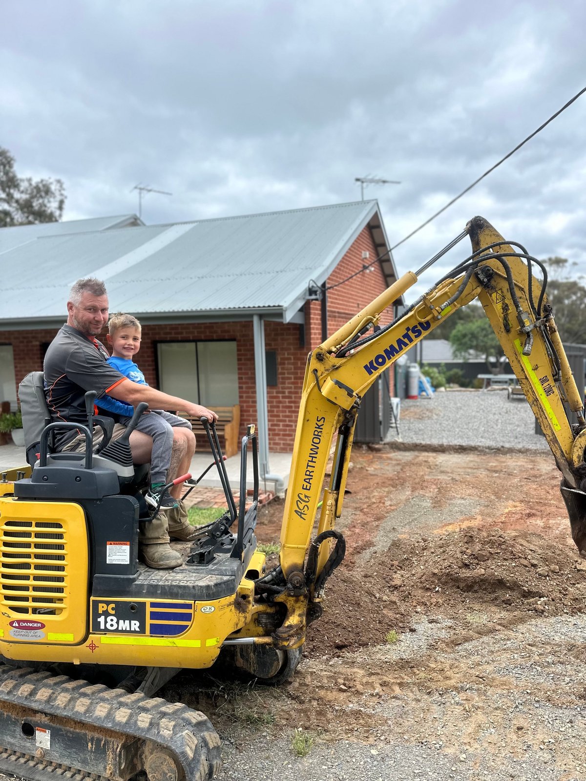 Adult and child sitting in the cab of a yellow mini excavator at a residential construction site