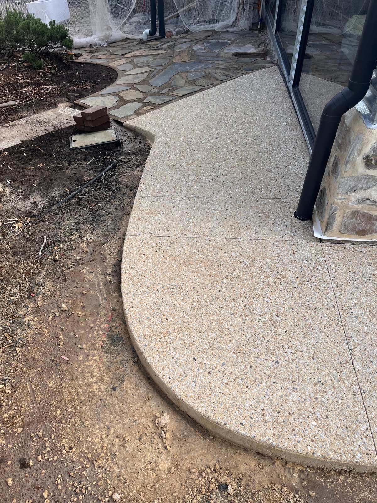Curved concrete patio leading to house entrance with landscaping and dark downspout visible