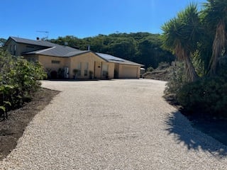 Single-story house with metal roof on gravel driveway surrounded by palm trees and green hillside