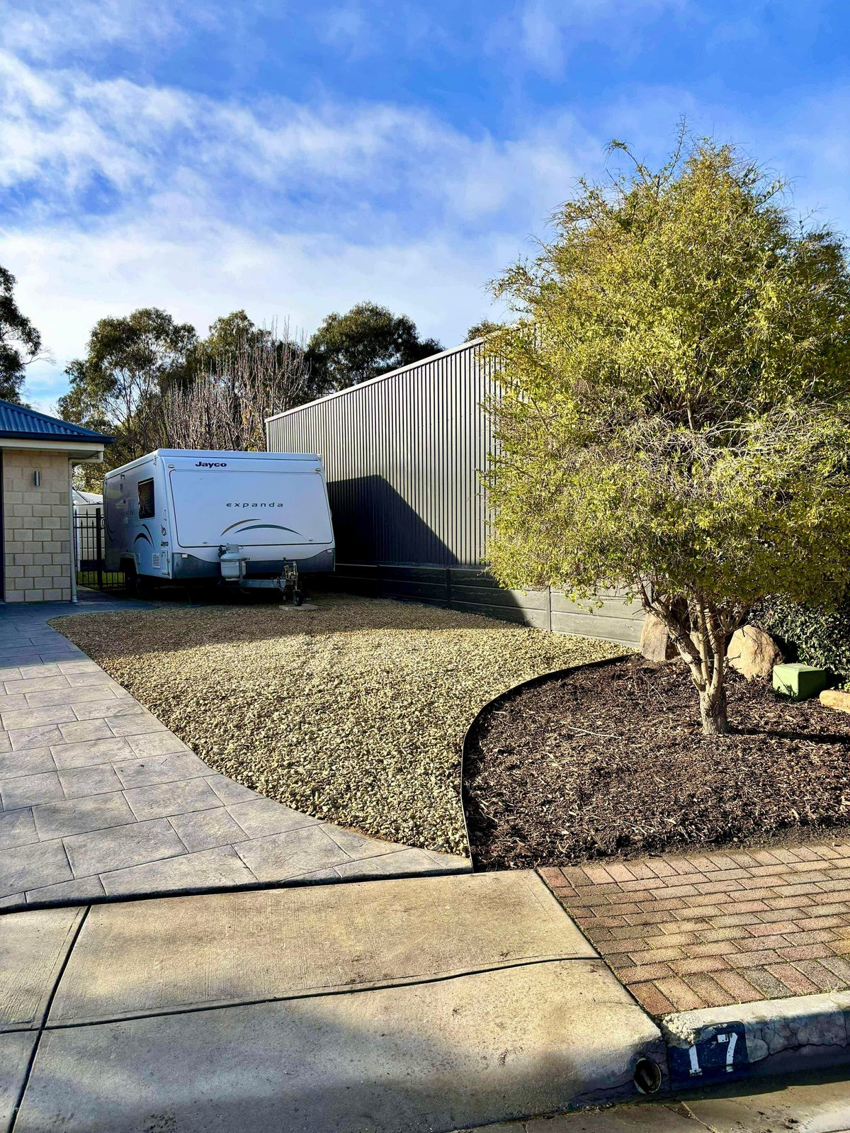 White travel trailer parked in driveway next to metal shed with landscaping and trees on clear sunny day