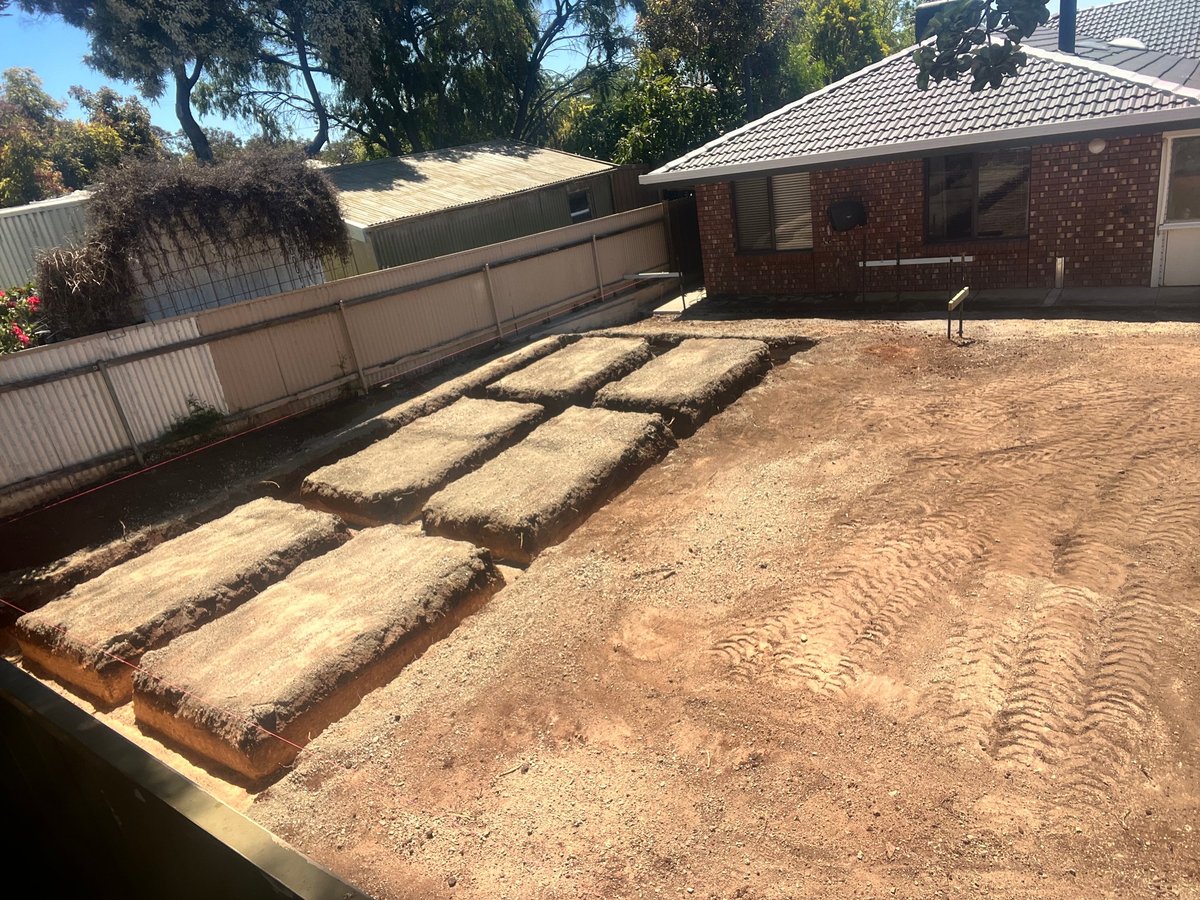 Residential driveway under construction with wooden forms laid out on dirt, brick house, and metal fence in background