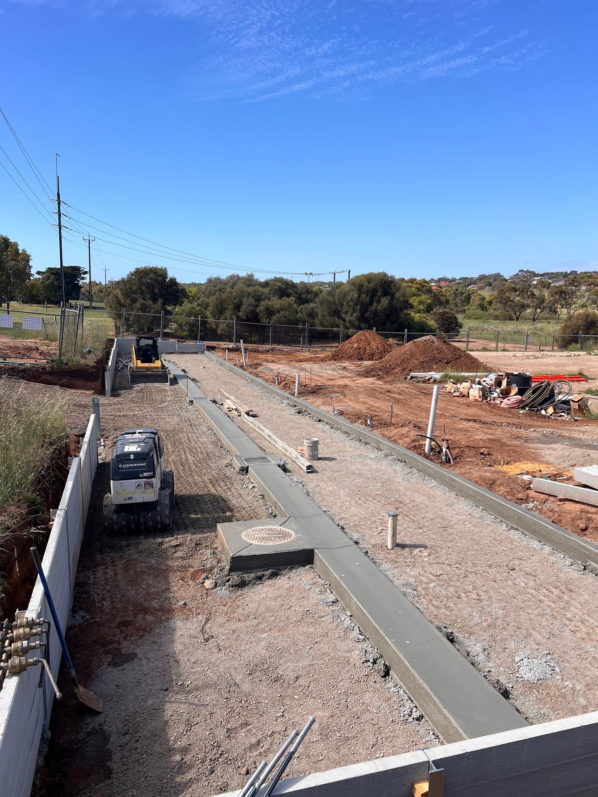 Construction site with freshly poured concrete foundation, machinery, and red dirt mounds under clear blue sky