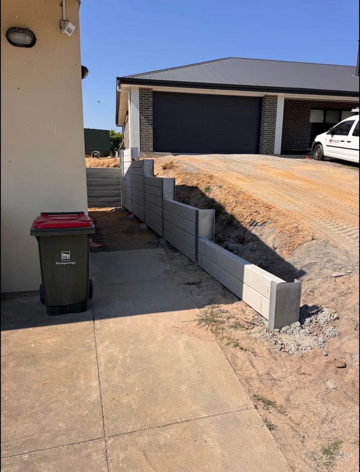Residential driveway with garbage bin in foreground, new brick garage with black doors, dirt landscaping, and white vehicle parked nearby