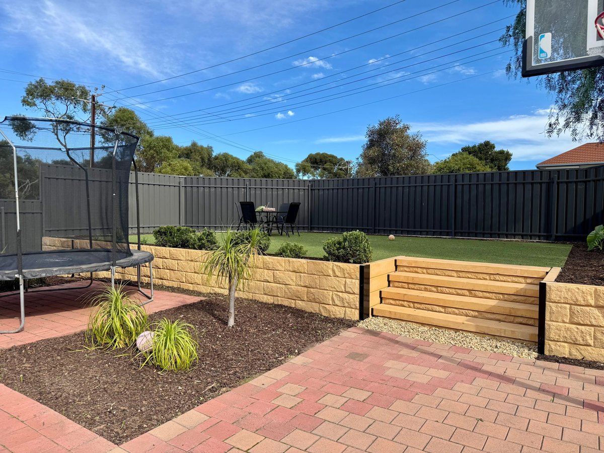 Suburban backyard with trampoline, wooden raised garden beds, green lawn, and black fencing under clear blue sky