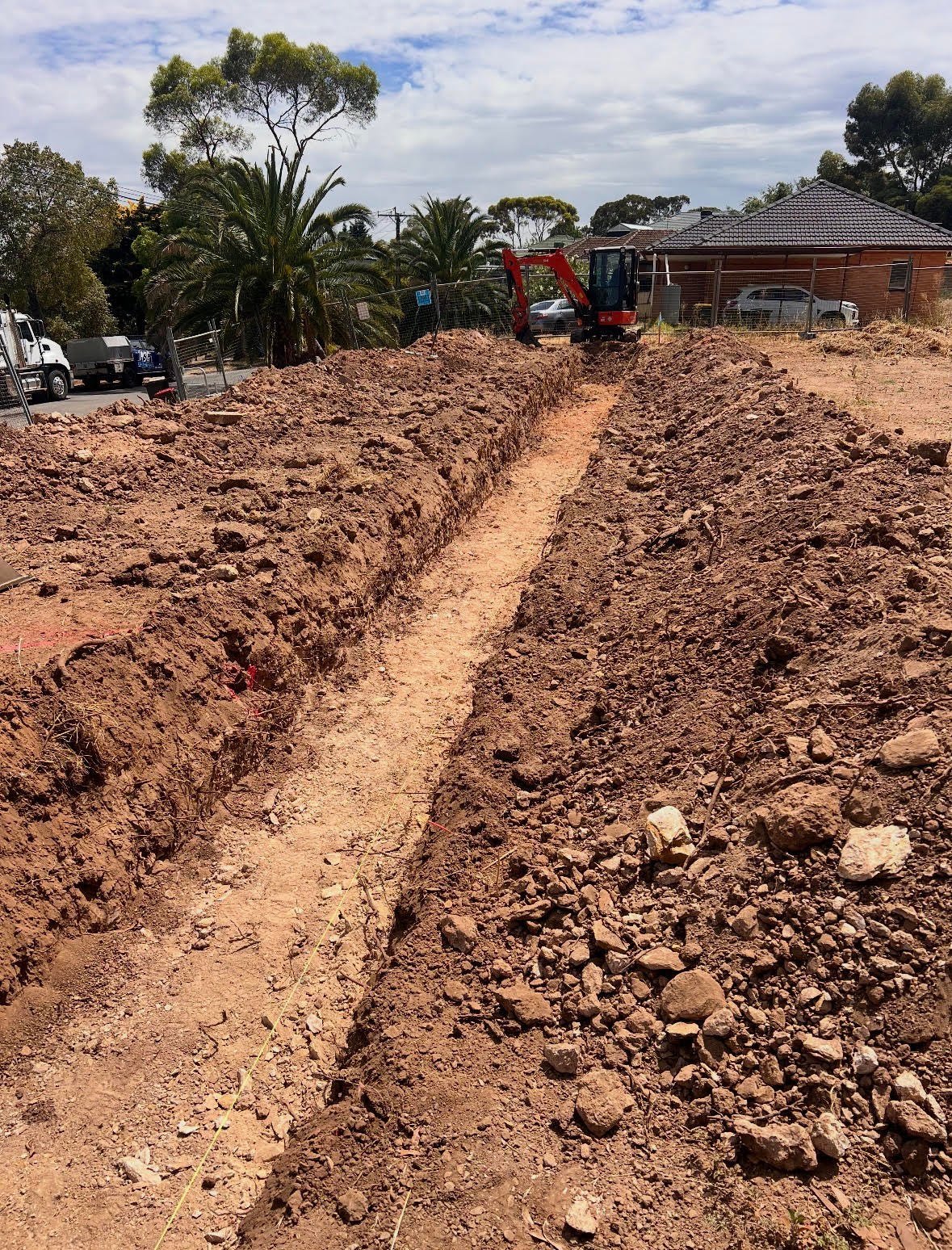 Excavated trench with dirt piles at residential construction site, palm trees and brick building visible in background