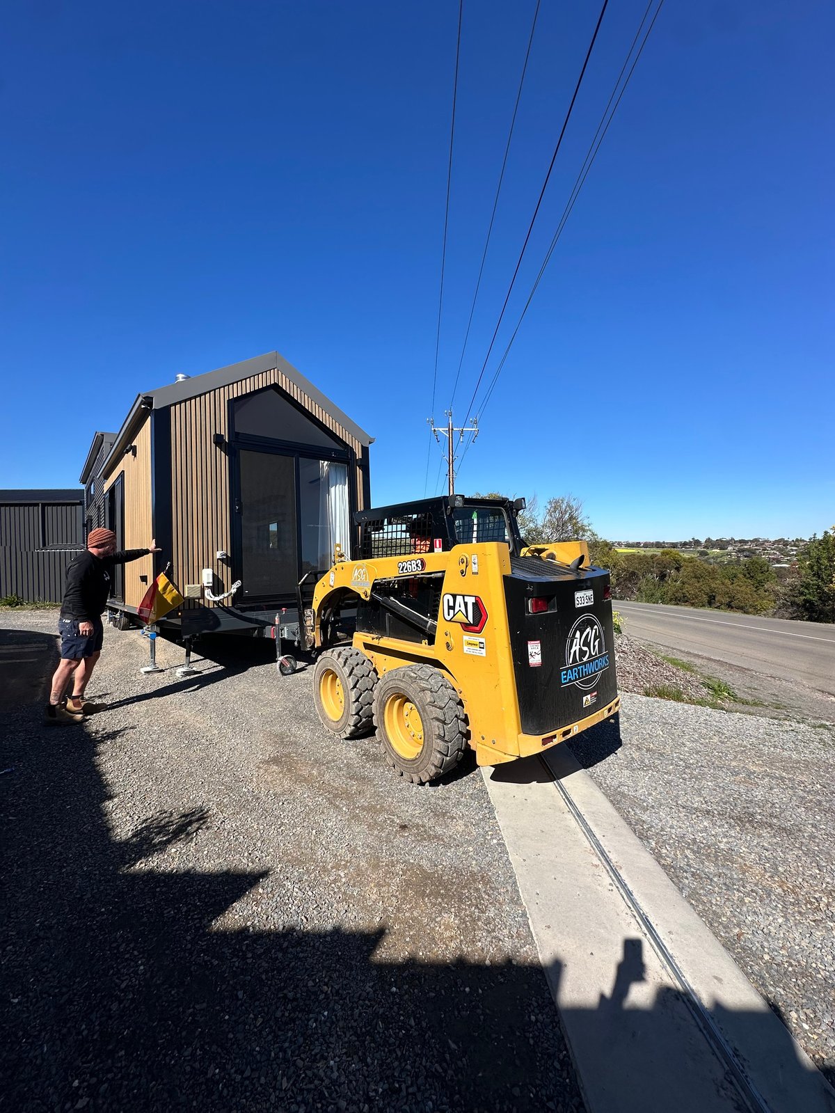 Yellow CAT skid steer loader positioning a metal shed on a rural road under clear blue sky