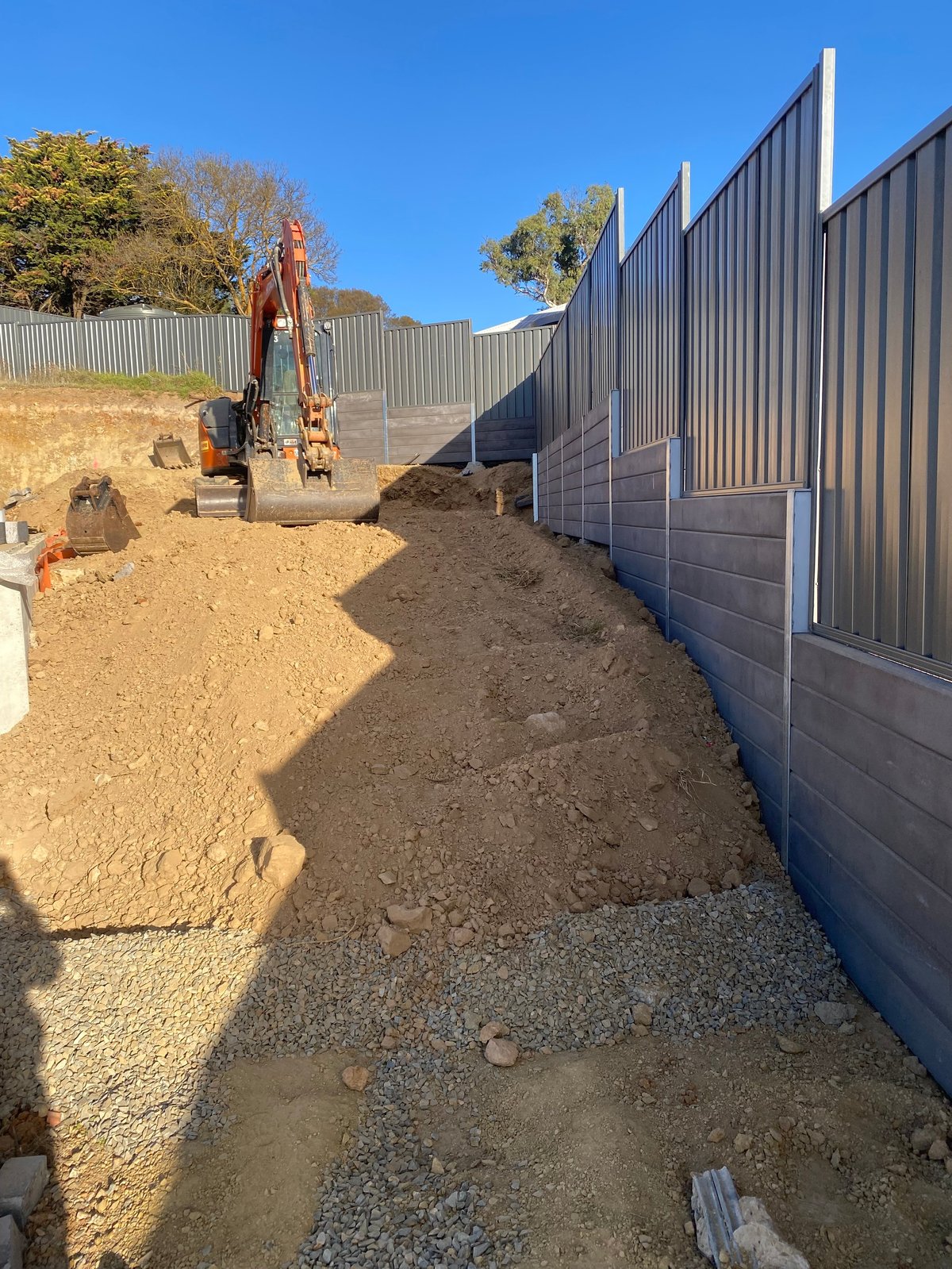 Excavator on dirt pathway between tall metal fencing with autumn trees in background under clear blue sky