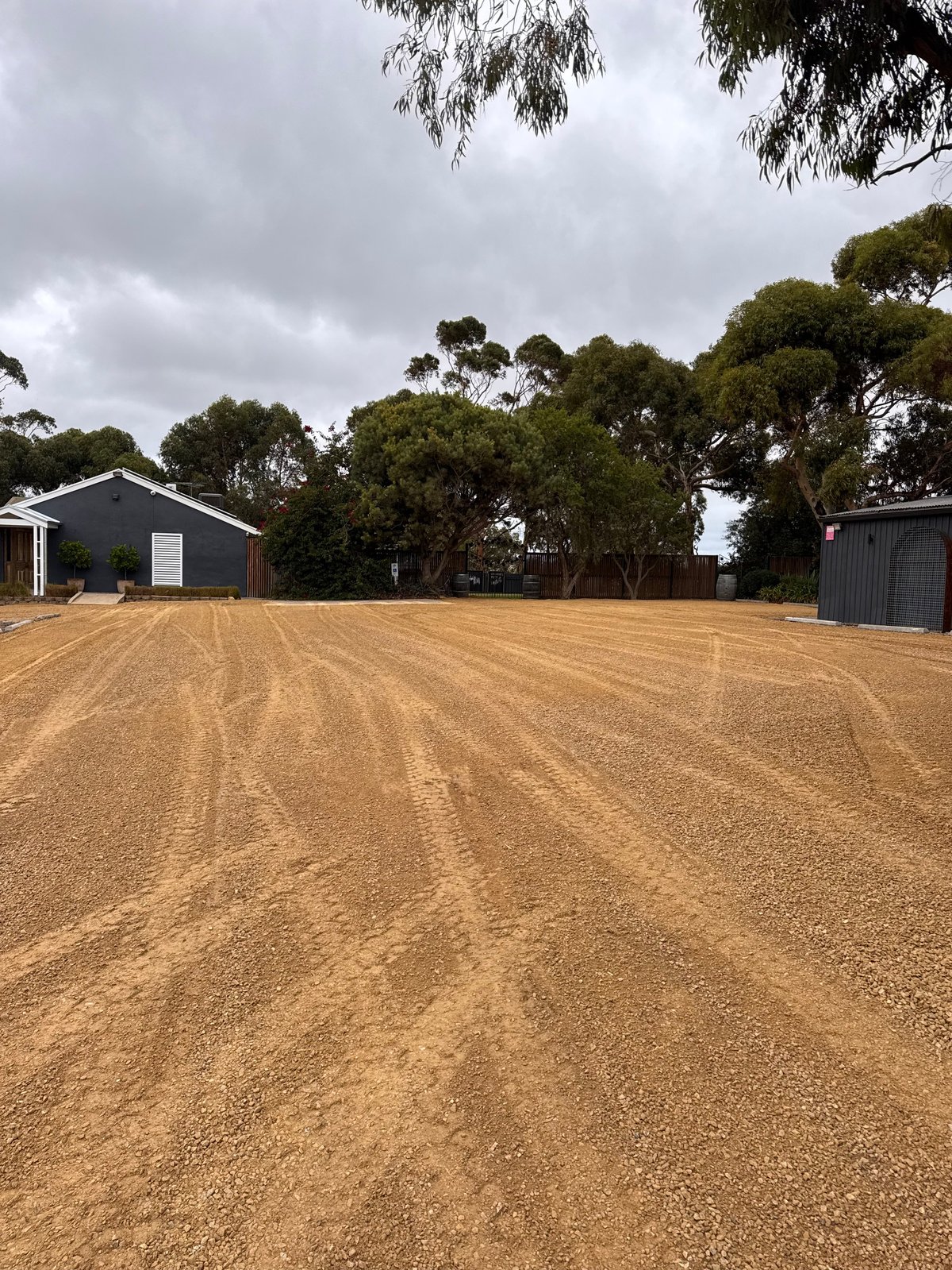 Rural property with cleared dirt driveway, dark farm buildings, eucalyptus trees, and overcast sky