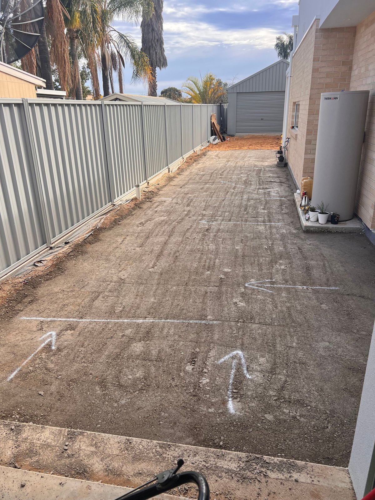 Outdoor residential driveway under construction with corrugated metal fence, brick building, and concrete being laid