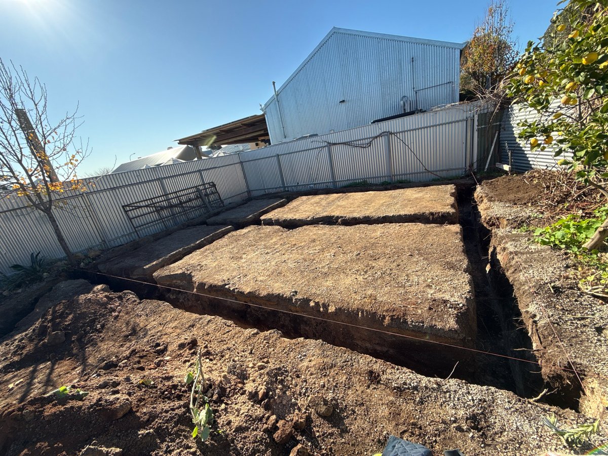Corrugated metal agricultural building with attached wooden fence enclosure on bare dirt ground under clear sky