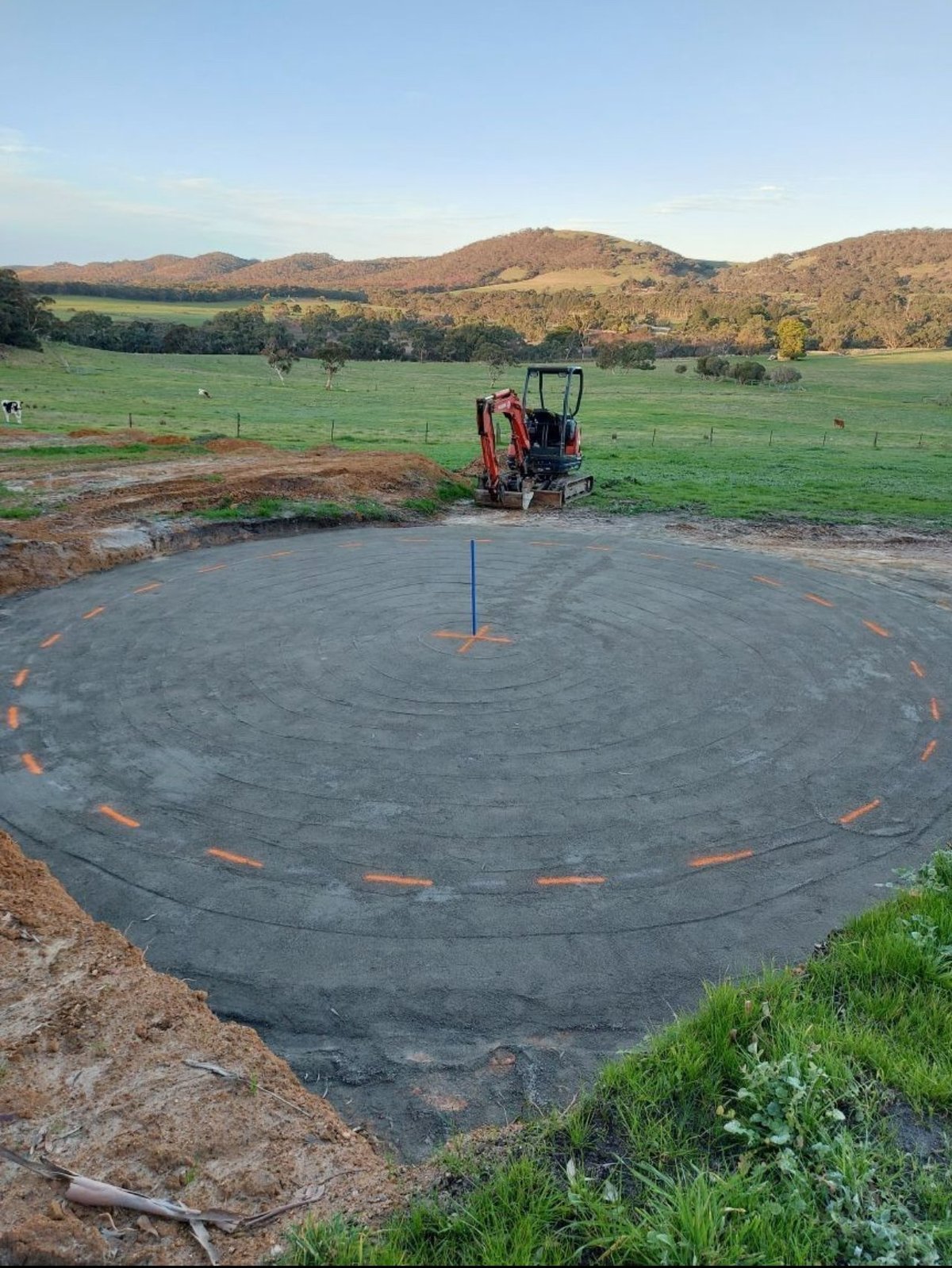 Circular farm pond with orange marker posts under construction, red machinery in background, green pasture and forested hills beyond