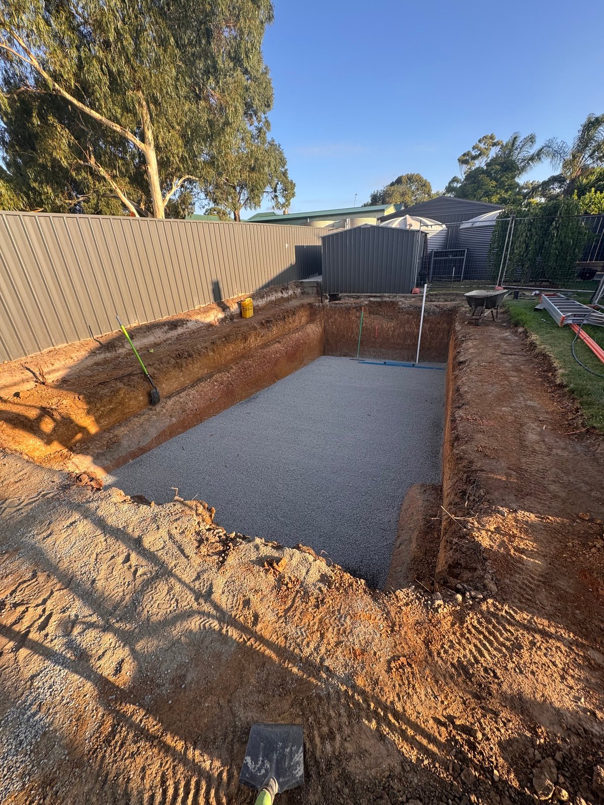 Excavated rectangular foundation pit with concrete base in rural backyard, surrounded by metal fencing and corrugated structures