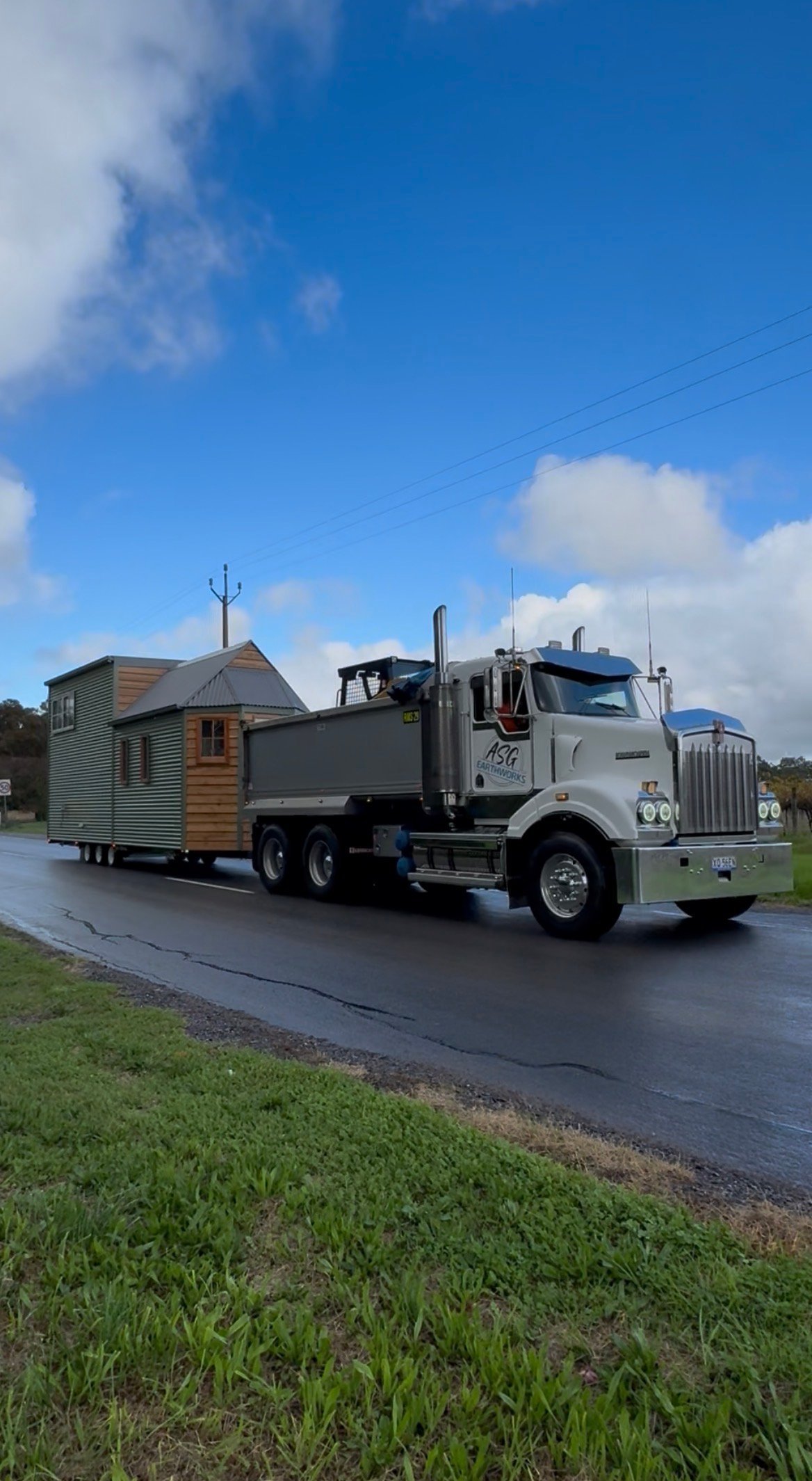 White semi-truck hauling a tiny house on a trailer down a wet paved road under blue sky