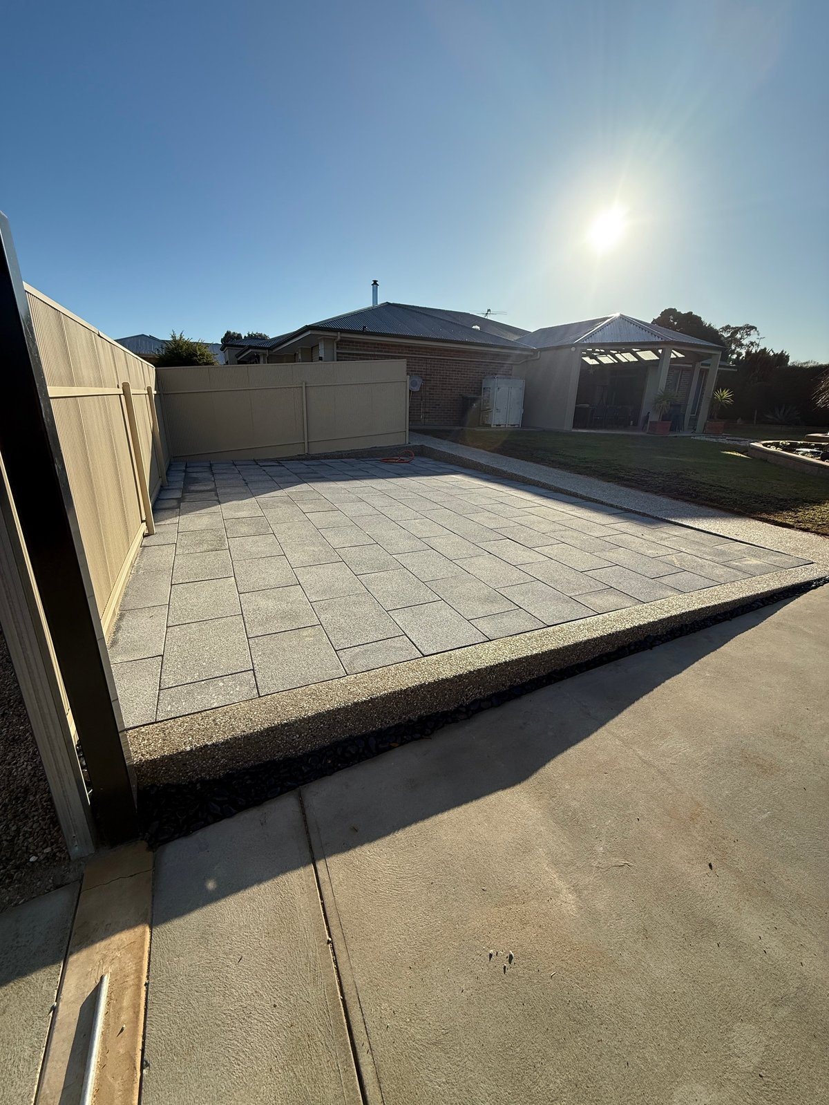 Modern paved courtyard with large tile flooring, beige fence, residential buildings with pergola, and clear blue sky