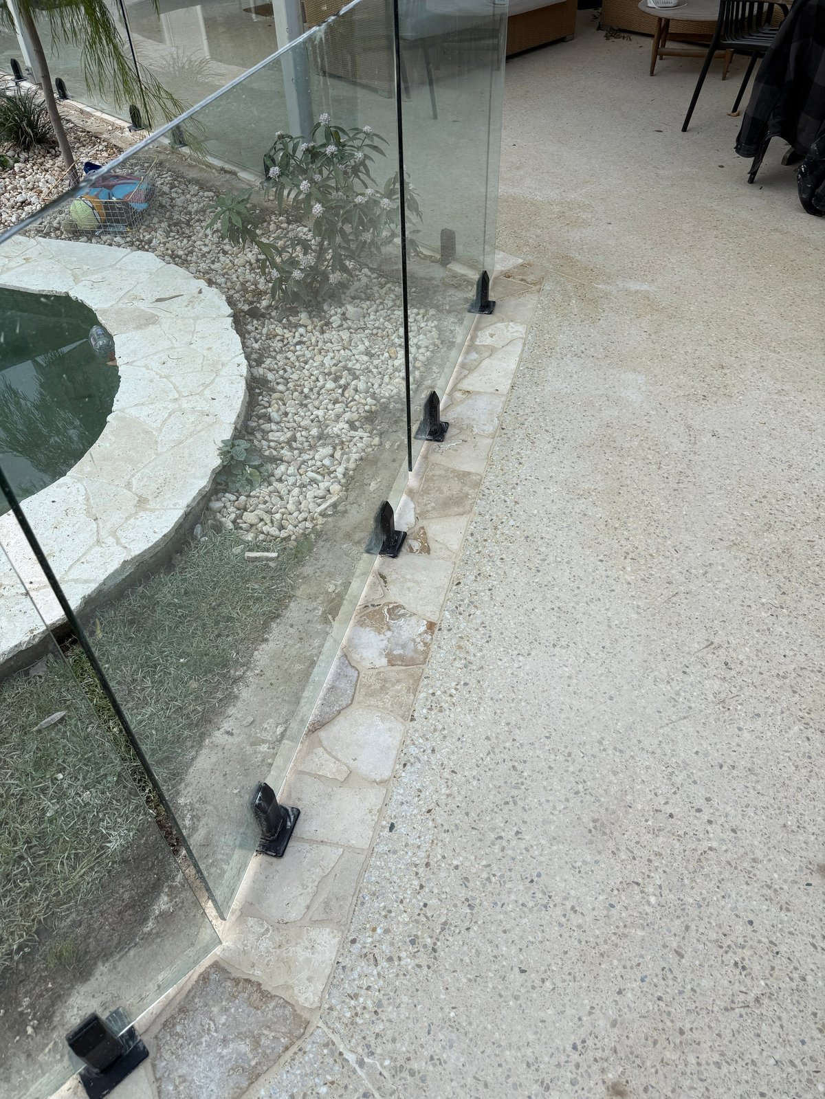 Glass panel barrier with black clamps separating indoor and outdoor spaces near landscaping and gravel area