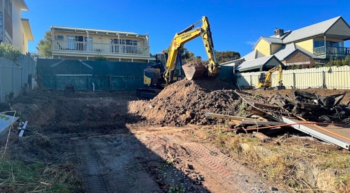 Yellow excavator operating at residential construction site with piles of dirt and debris under clear blue sky
