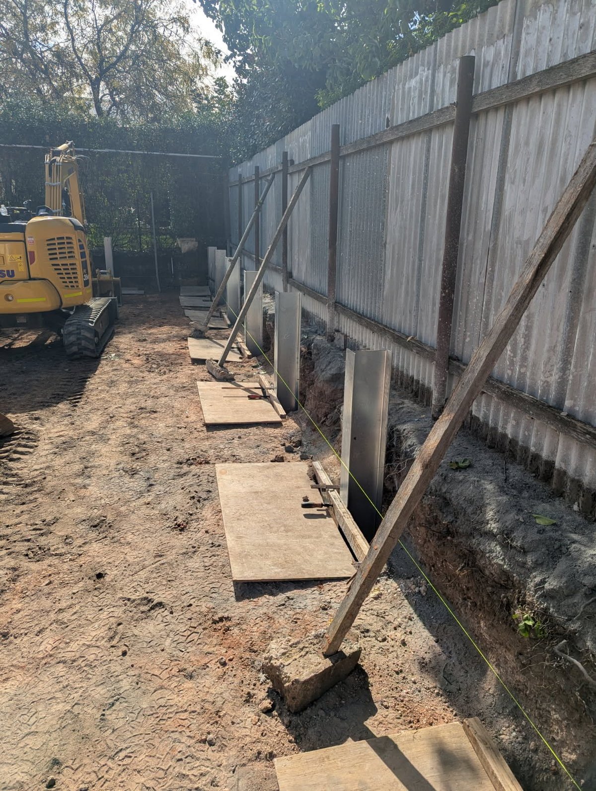 Construction site with corrugated metal fence, wooden platforms, and yellow excavator equipment on dirt ground