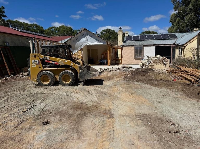 Yellow CAT skid steer loader on dirt lot with brick house under renovation and construction materials
