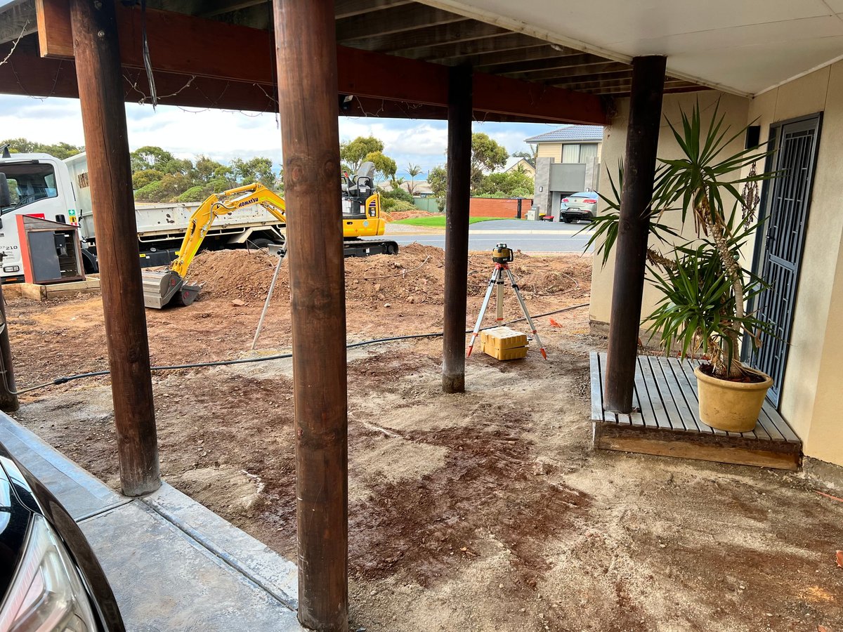 Construction site under a covered structure with wooden posts, potted plants, and yellow excavator visible in background dirt yard