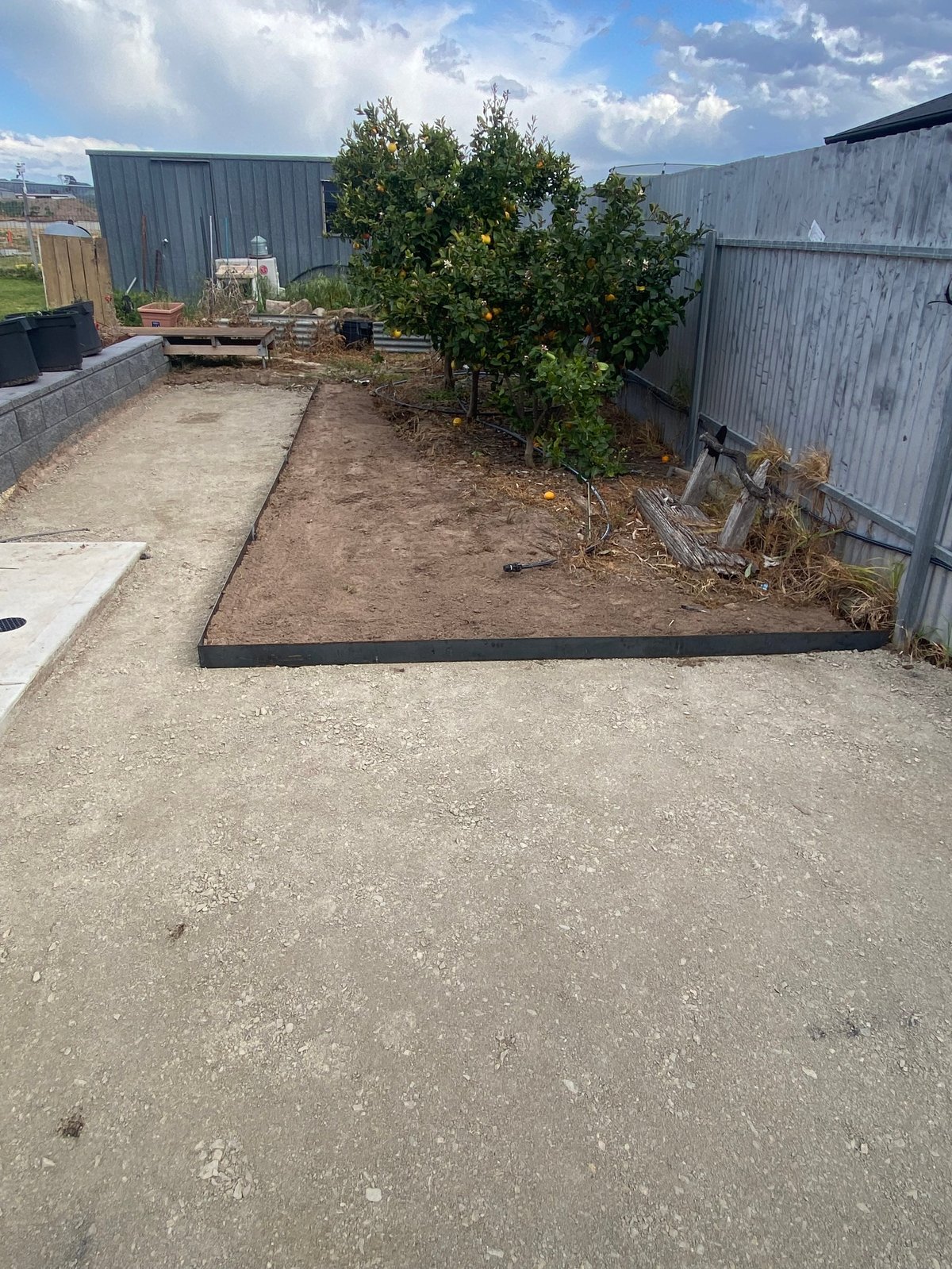 Industrial yard with raised garden bed, metal buildings, trees, and concrete ground under cloudy sky