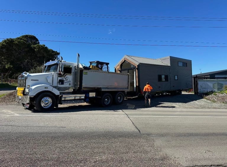 Large white semi-truck with flatbed trailer carrying a black modular building, parked on a paved road with a worker in orange visible nearby