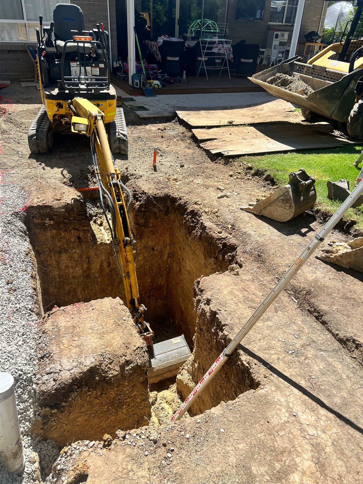 Yellow excavator digging a trench in a residential yard with construction equipment and garage visible