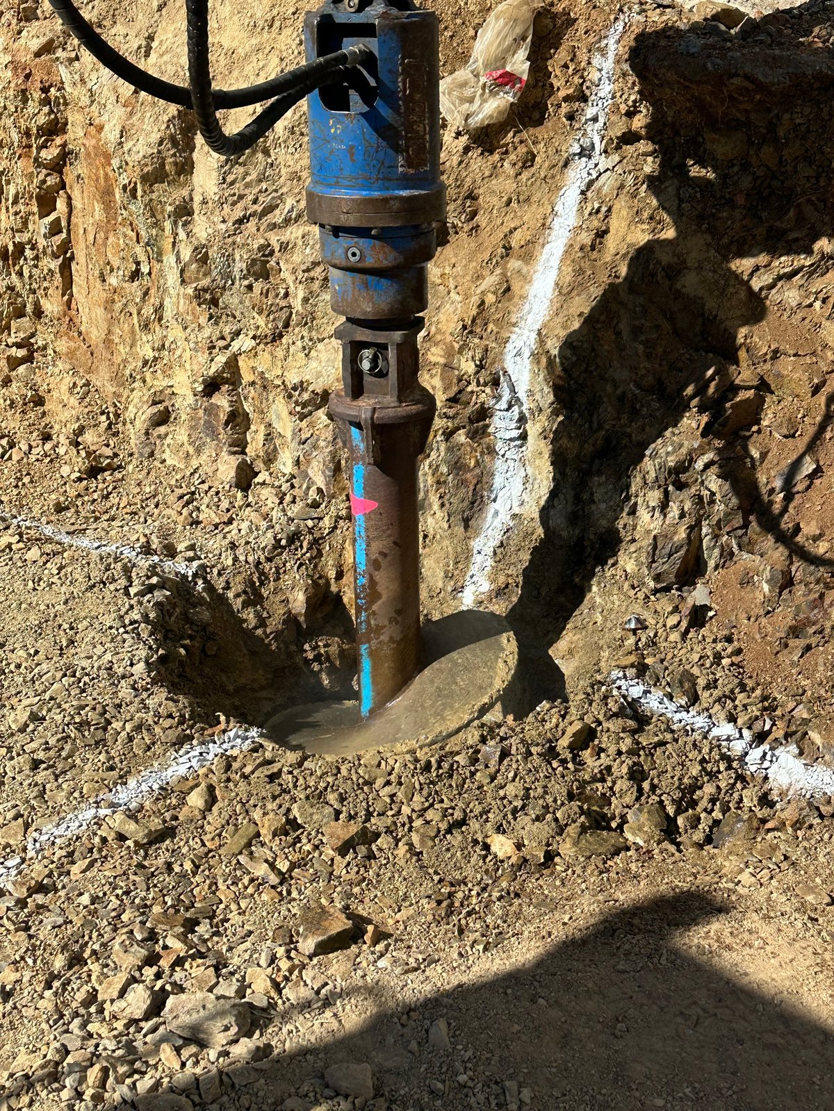Blue jackhammer breaking concrete in excavated dirt, mounted on pneumatic arm at construction site