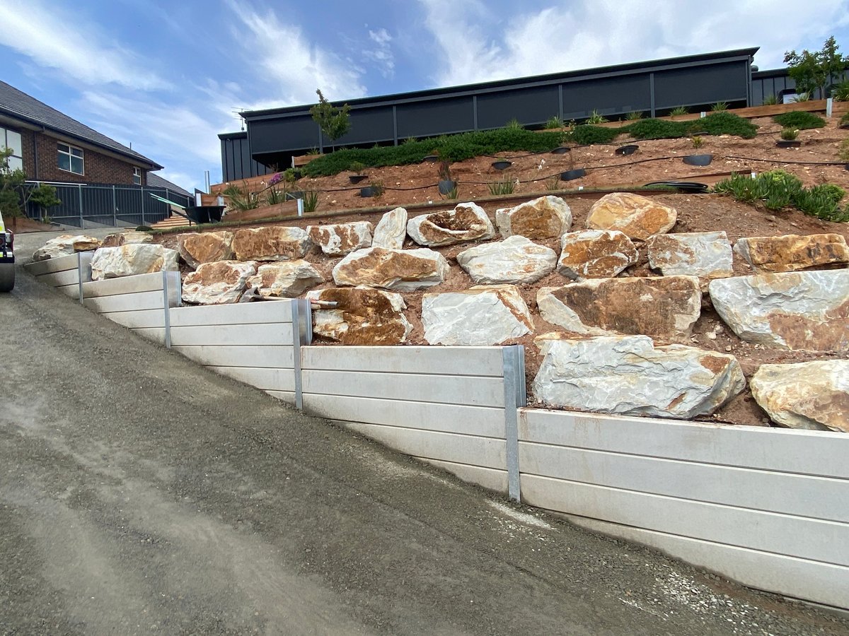 White metal retaining wall holding large boulders on a hillside with buildings and green vegetation visible above