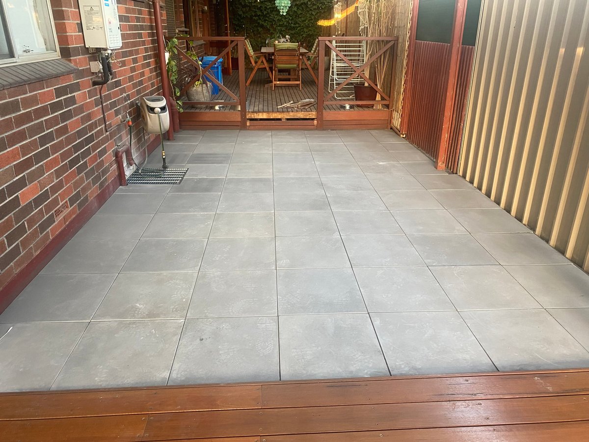 Paved patio area with gray tiles, wooden deck border, brick wall, and corrugated metal fencing on sides, with potted plants and furniture in background