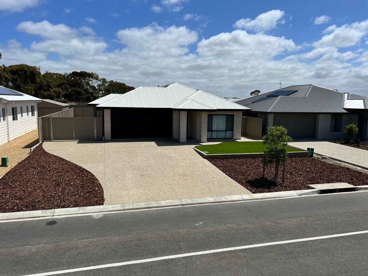 Modern single-story home with beige exterior, dark roof, concrete driveway, and landscaped front yard with mulch and a young tree