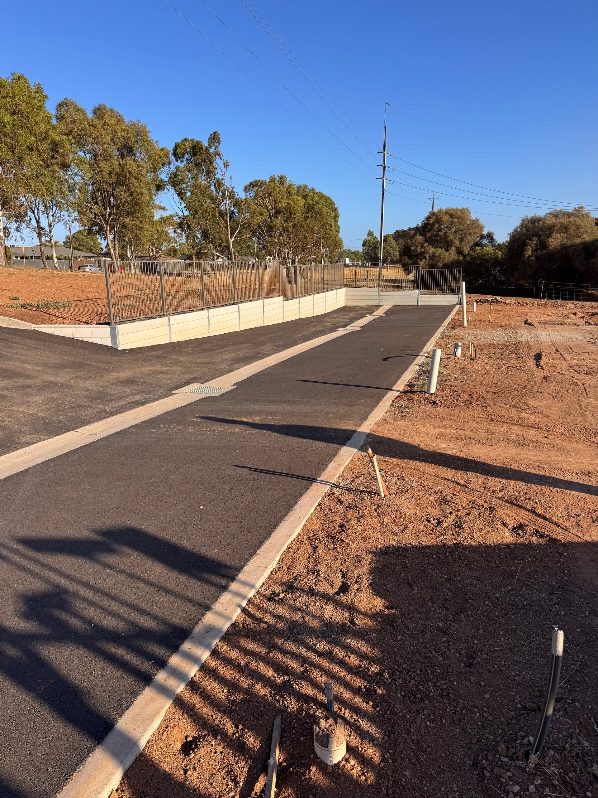 New asphalt pathway with white edge lines in a recreational park with metal fencing, red dirt, and eucalyptus trees under clear blue sky