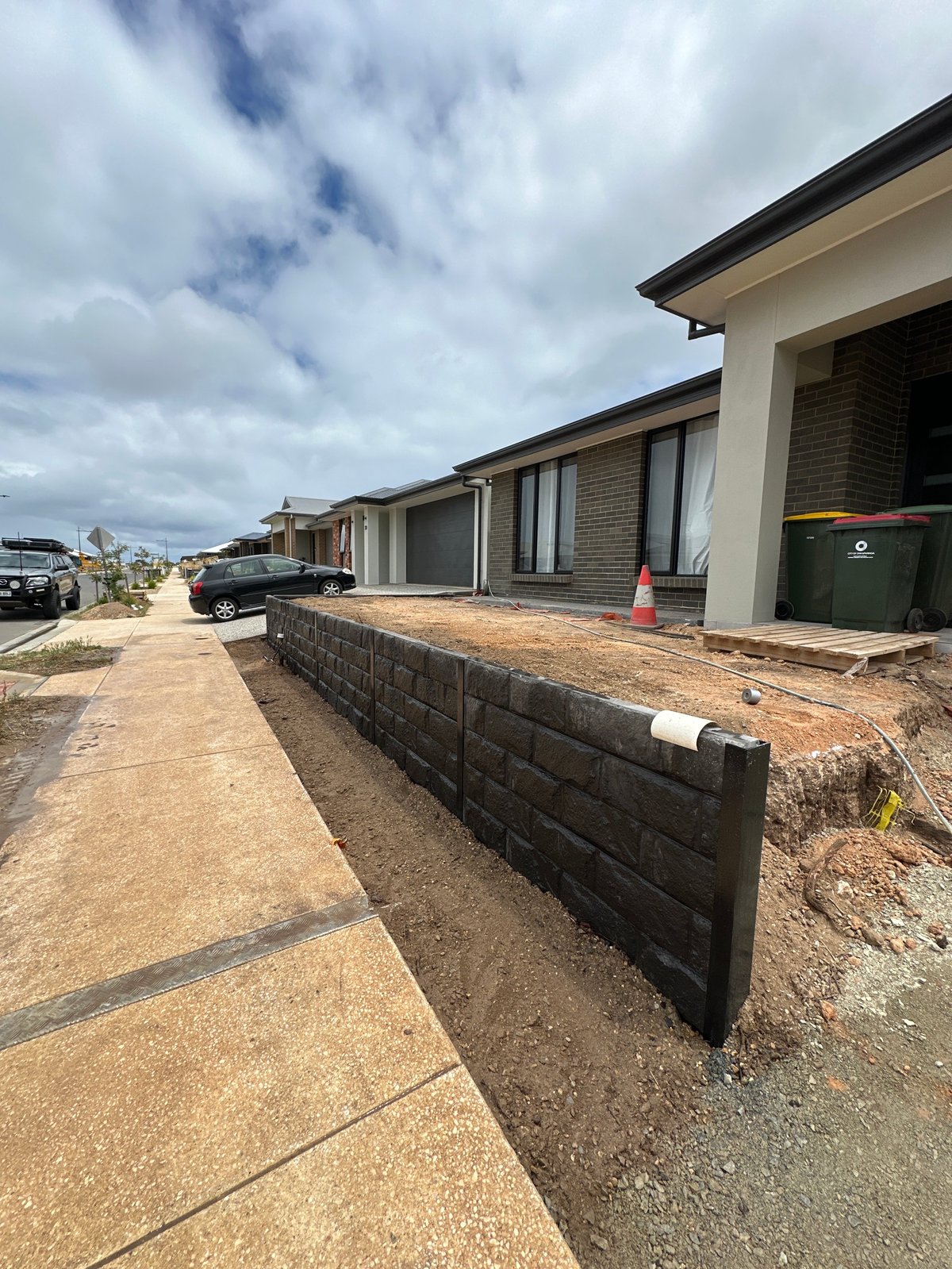 Modern residential home with brick and render exterior, featuring a dark retaining wall along the front pathway, parked cars visible on the street, and blue cloudy sky