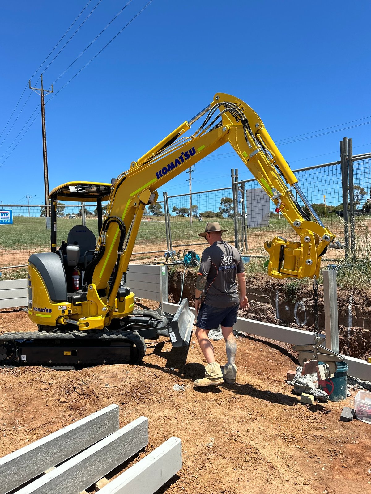 Person operating a yellow Komatsu excavator on a dirt construction site with metal fencing and wooden materials visible