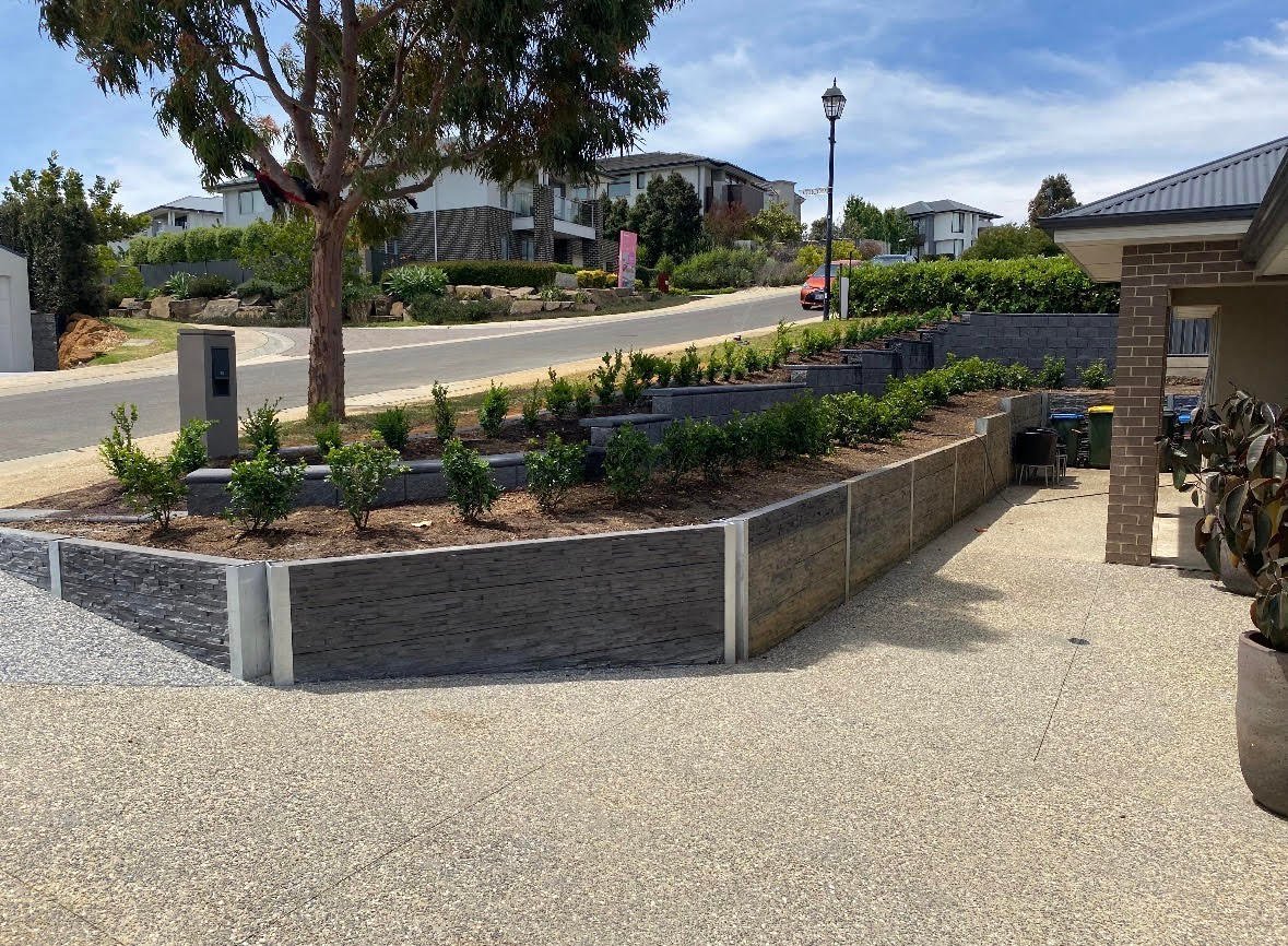 Residential driveway with curved concrete planters containing plants and trees, adjacent street, and brick wall on right