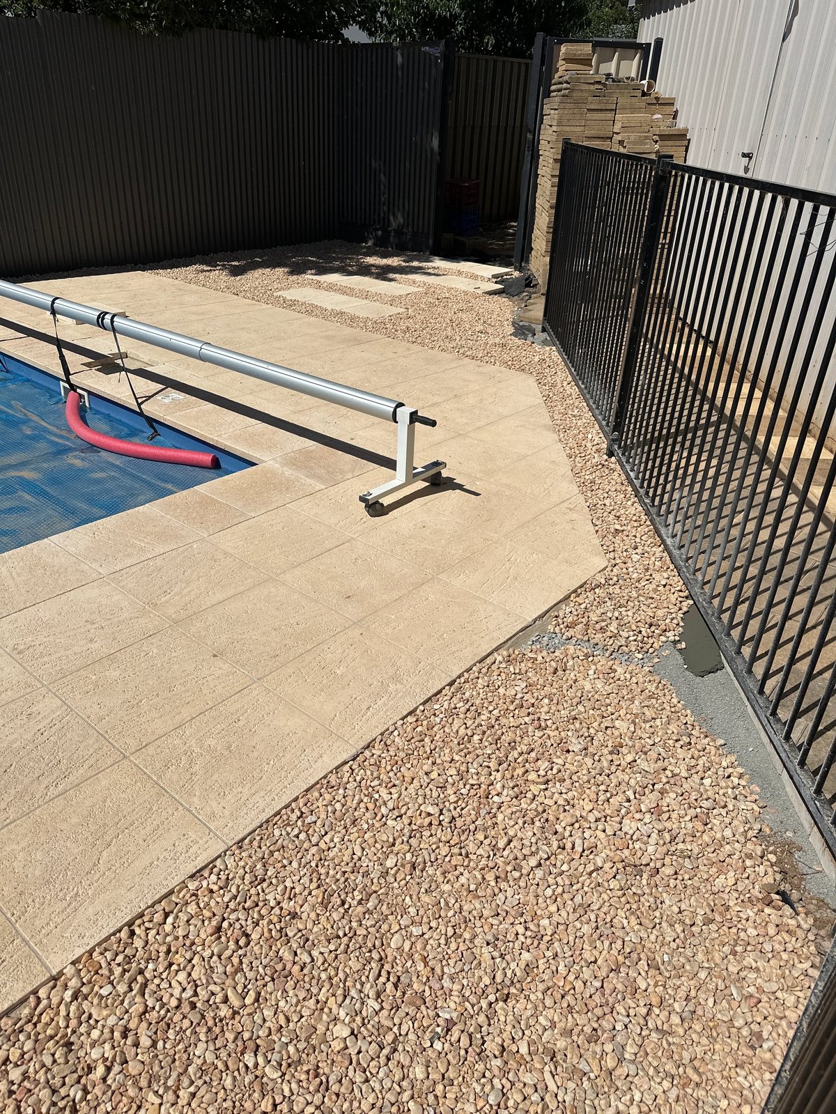 Backyard with swimming pool, metal railing, paved patio, gravel ground, and black metal fence against corrugated background
