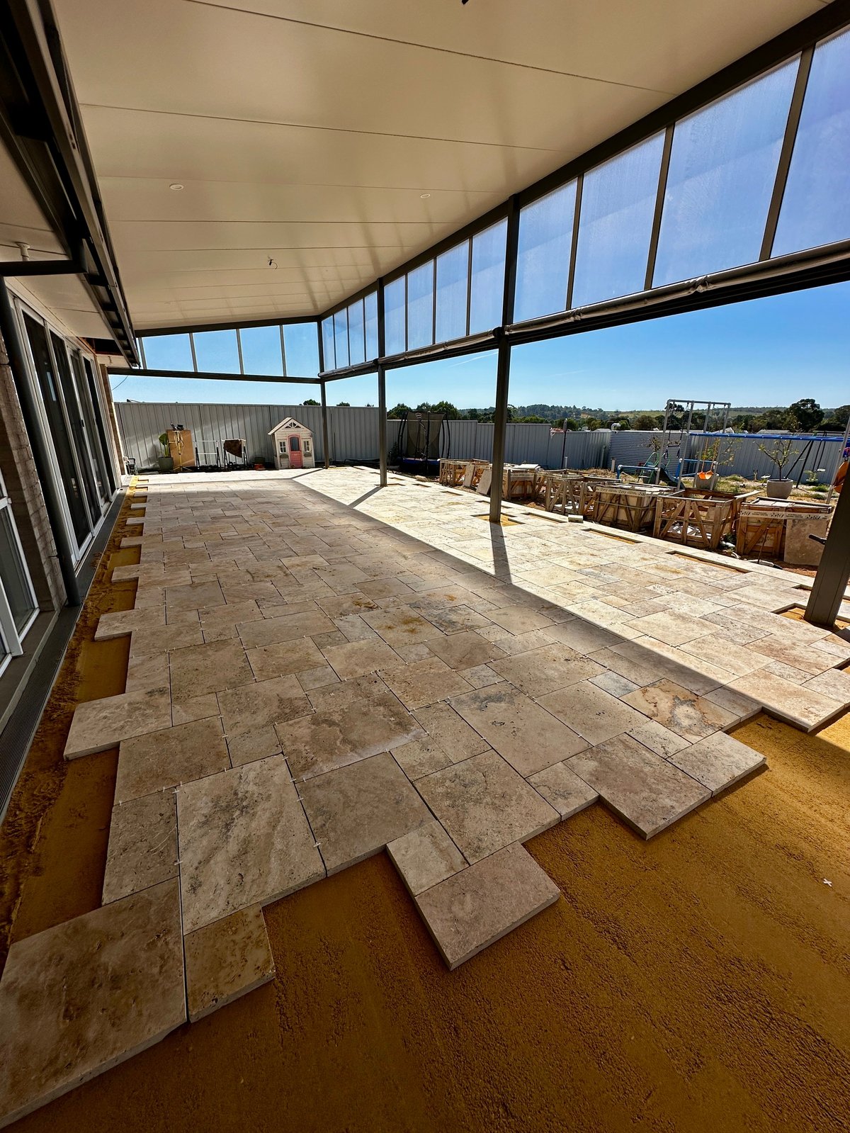 Sunlit covered patio with large windows overlooking a waterfront lake, showing pavers on the floor and distant trees