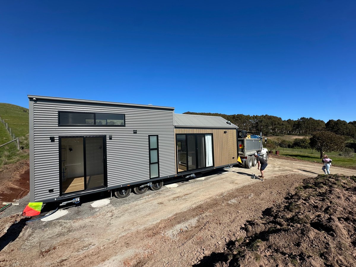 Modern tiny house on wheels with corrugated metal siding in rural setting with people standing nearby and clear blue sky