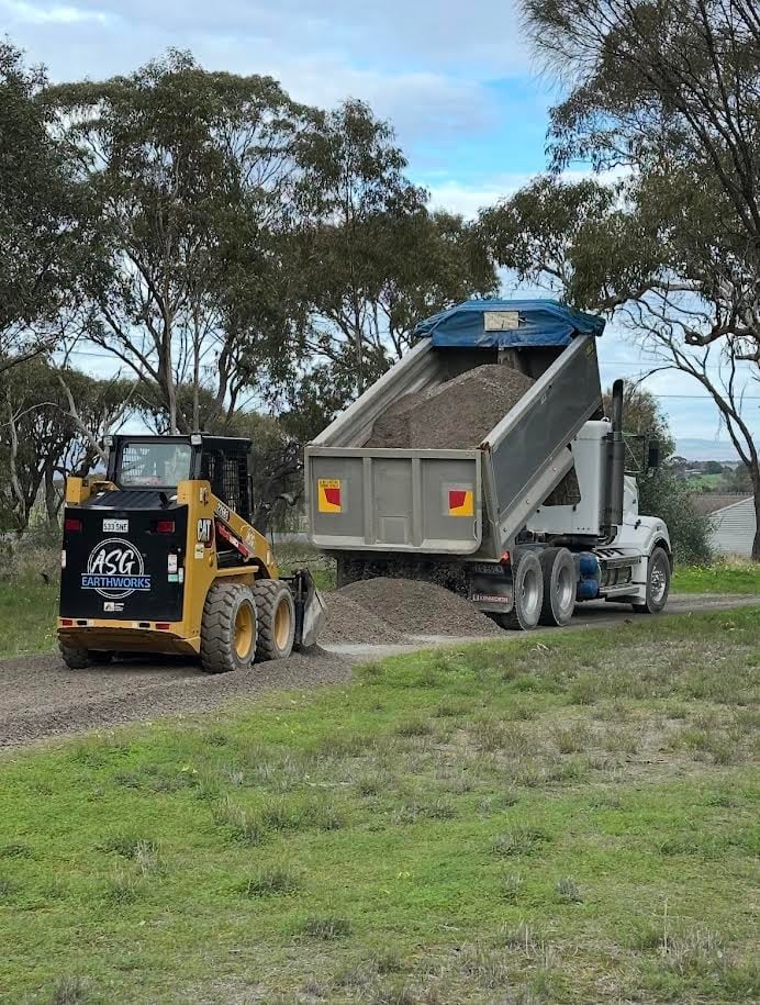 Yellow skid steer loader loading gravel into a gray dump truck bed on a rural road with eucalyptus trees